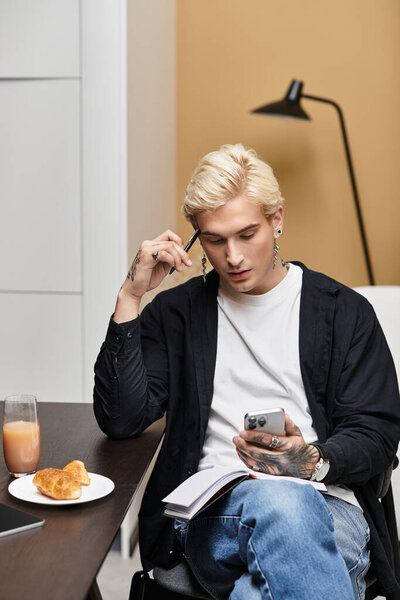 Красивый молодой человек сидит в уютной квартире, глубоко думая, держа ручку и телефон.