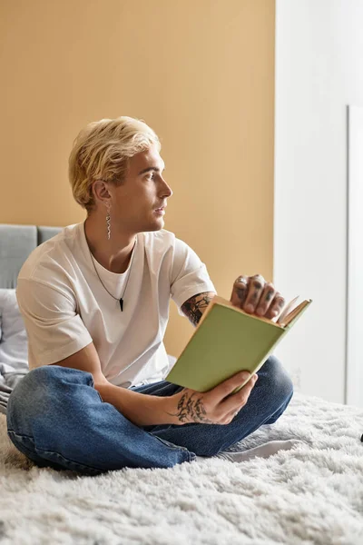 Dövmeleri olan yakışıklı genç adam yumuşak bir halıda bacak bacak üstüne atarak oturuyor ve evinde kitap okuyor..