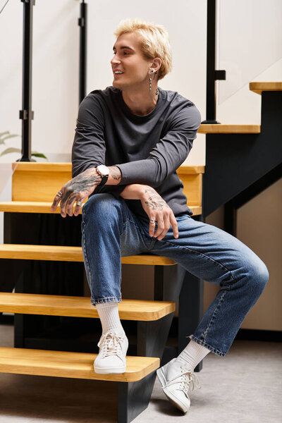 Красивый молодой человек удобно сидит на деревянных ступеньках, демонстрируя свой повседневный стиль и татуировки.