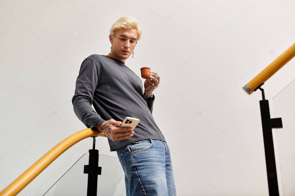 A stylish young man relaxes with a cup in hand, focused on his phone in a contemporary setting.