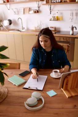 Bir kadın evdeki serbest işlerini yönetir, kahveden zevk alır ve parlak bir mutfakta notlar yazar..