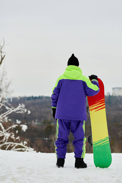 A man in colorful outerwear prepares to snowboard down a snowy slope on a winter day.