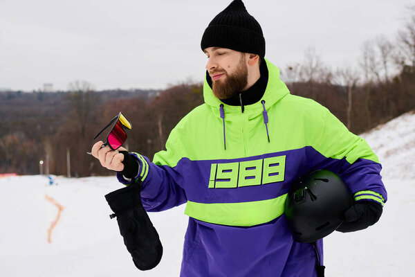 Бородатый человек держит красочные очки во время сноуборда на снежном склоне в окружении зимних пейзажей.