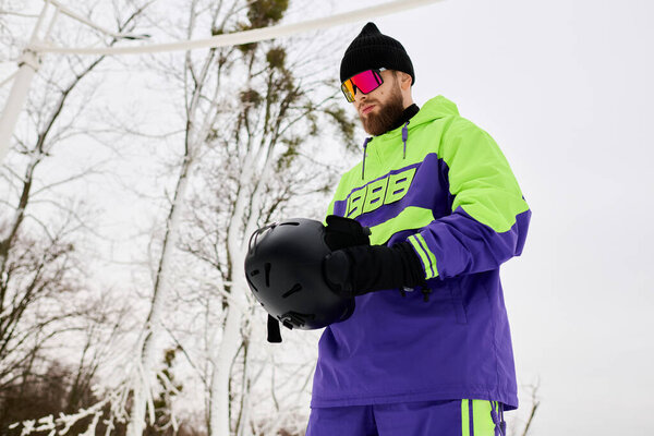 На снежном склоне бородатый мужчина держит свой шлем для сноуборда, готовый к захватывающей поездке.