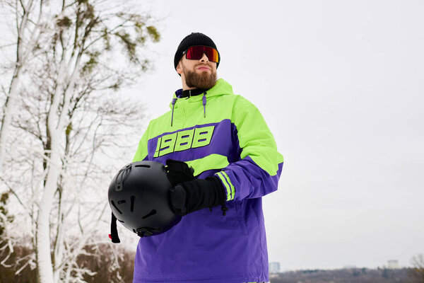 Бородач стоит на снежном склоне, готовясь к сноуборду в разноцветной зимней одежде.