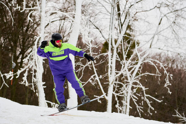 Человек с бородой умело катается на сноуборде по снежному склону в окружении деревьев.