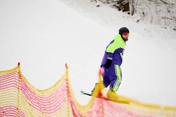 A bearded man skillfully snowboards down a snowy slope wearing bright outerwear.