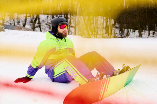 Snowboarder with a beard rests on the snowy slope, enjoying the winter sports atmosphere.