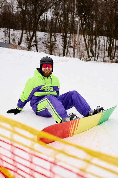 Бородатый мужчина в разноцветной верхней одежде сидит на снежном склоне, улыбаясь после сноуборда.