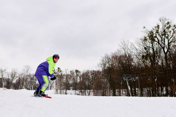 Бородатый человек умело катается на сноуборде по снежному склону, наслаждаясь зимним днем среди высоких деревьев.