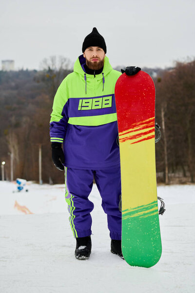 Бородатый мужчина гордо стоит на снежном склоне со своим красочным сноубордом, готовый к поездке.