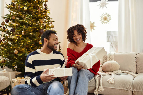 Радостная афро-американская пара разворачивает подарки у своей красиво украшенной елки.