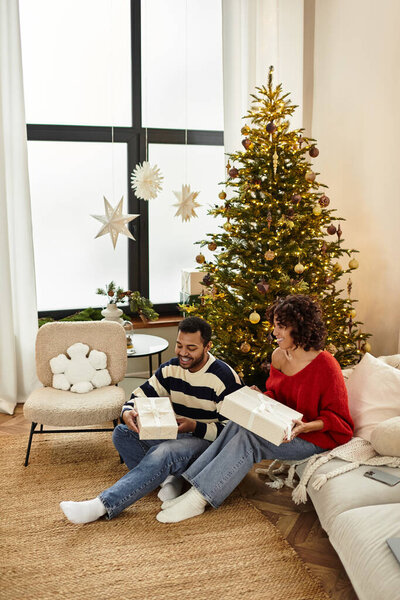Радостная афроамериканская пара разворачивает подарки своей красиво украшенной елкой.