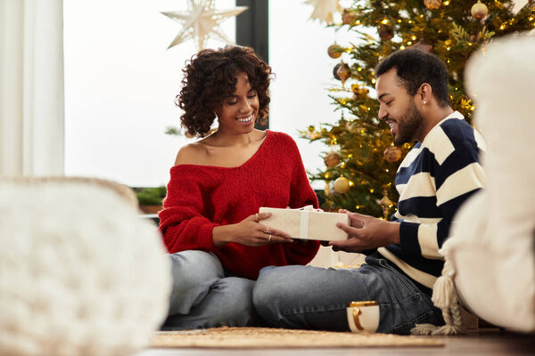 Радостная афроамериканская пара делится подарками в своем стильно оформленном рождественском доме.