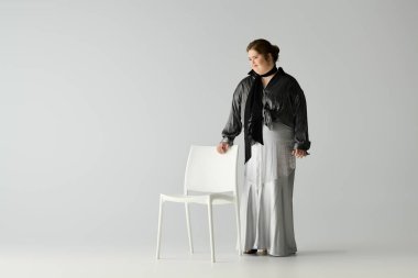 Genç bir kadın beyaz bir sandalyenin yanında durarak özgünlüğünü zarafetle ifade eder..