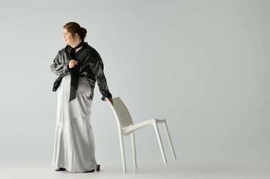 Down sendromlu genç bir kadın şık bir stüdyoda sandalyeyle gururla durur ve mutluluk saçar..