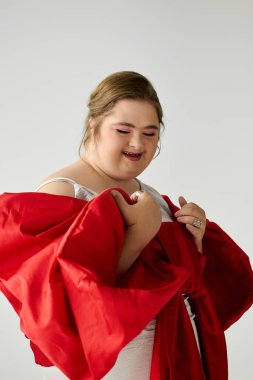 Down sendromlu genç bir kadın elbisesinin yanında neşe içinde kırmızı kumaşı tutarken gülümsüyor..