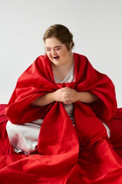 Down sendromlu genç bir kadın, kırmızı bir battaniyeyi kucaklarken nazikçe oturuyor ve gülümsüyor..