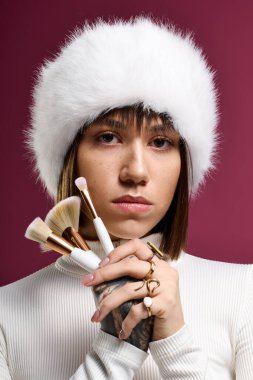 Moda kıyafetli genç güzel cesur pembe bir arka plana karşı makyaj fırçalarını sergiliyor.