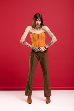 Kalın turuncu korseli ve şık pantolonlu kendine güvenen bir kadın kırmızı zeminde zarifçe poz veriyor..