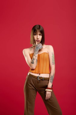 Dövmeleri olan genç bir kadın kırmızı bir arkaplanda şık bir şekilde poz veriyor..