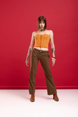 Zarif genç bir kadın kendine güvenerek ayakta duruyor, parlak turuncu bir bluz ve kahverengi bir pantolon giyiyor..