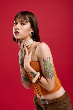 Şık bir genç kadın kendine güvenerek poz veriyor, dövmeleri ve şık kıyafetleri kırmızı arka planda sergiliyor..