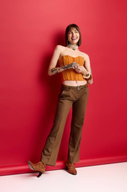 Parlak giyimli genç bir kadın şık kıyafetler içinde cesur kırmızı bir zemine karşı mutlu bir şekilde duruyor..