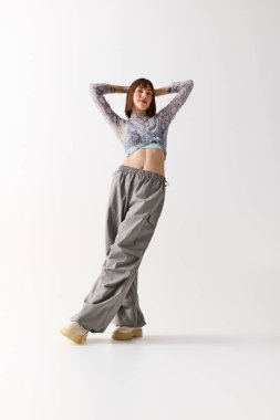 Genç kadın kendine güvenen bir şekilde poz veriyor. Parlak bir alanda elleri başının arkasında..