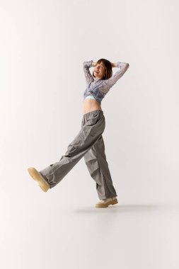 Genç bir kadın şık kıyafetlerini sergilerken gülümsüyor ve minimalist bir stüdyoda poz veriyor..