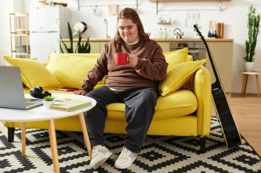 Down sendromlu neşeli genç bir kadın parlak sarı bir kanepede oturur ve evdeki içkisinin tadını çıkarır..