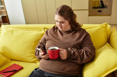 Down sendromlu genç bir kadın sarı bir koltukta rahatlıyor, kahvesini yudumluyor ve mesajlaşıyor..