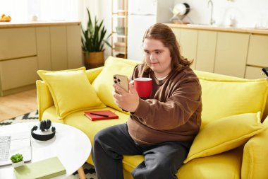 Down sendromlu genç bir kadın sarı bir koltukta dinlenirken akıllı telefonunu kullanıyor ve çayını yudumluyor..