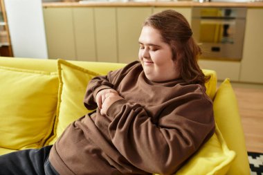 Down sendromlu genç bir kadın, parlak bir oturma odasındaki sarı bir kanepede mutlu bir şekilde oturur..