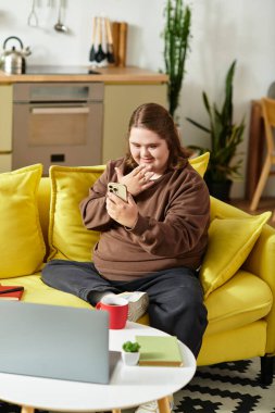 Down sendromlu genç bir kadın sarı bir kanepede akıllı telefonunu kullanırken gülümsüyor..