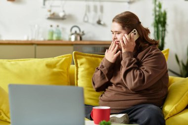 Down sendromlu genç bir kadın açık sarı bir kanepede oturup dinlenirken telefonla konuşuyor..