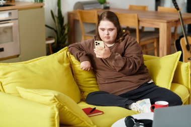 Down sendromlu genç bir kadın, akıllı telefonuyla meşgul sarı bir kanepede dinleniyor..