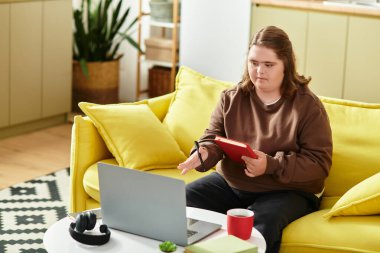 Down sendromlu bir kadın parlak sarı bir koltukta dizüstü bilgisayarıyla ve defteriyle meşgul olarak rahatlıyor..