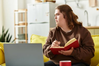 Down sendromlu genç bir kadın kanepede oturur, evdeki bir deftere yazar, düşünceli görünür..