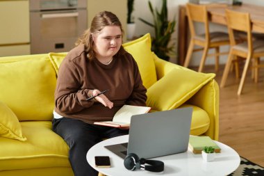 Down sendromlu genç bir kadın evdeki sarı koltukta oturur ve defterine odaklanır..