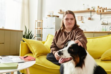 Down sendromlu genç bir kadın köpeğini evde okşarken parlak bir kanepede dinleniyor..