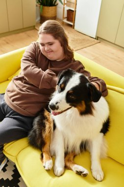 Down sendromlu genç bir kadın sarı bir koltukta oturuyor, köpeğine sarılırken gülümsüyor..