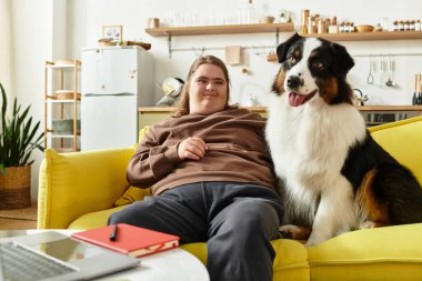 Evdeki sarı kanepede dinlenen genç bir kadın, mutlu köpeğinin yanında oturuyor ve gülümsüyor..