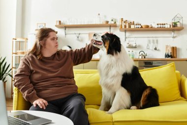 Down sendromlu genç bir kadın sarı bir kanepede rahatlıyor, köpeğini evde okşuyor..