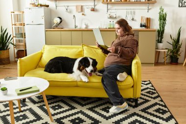 Down sendromlu genç bir kadın sarı bir kanepede rahatlar ve yanında köpeğiyle kitap okur..