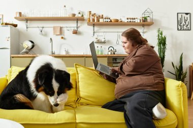 Genç bir kadın, köpeği yanında otururken dizüstü bilgisayarına odaklanmış parlak sarı bir kanepede dinleniyor..