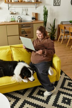 Down sendromlu genç bir kadın canlı sarı bir koltukta oturmuş köpeğiyle kitap okuyor..