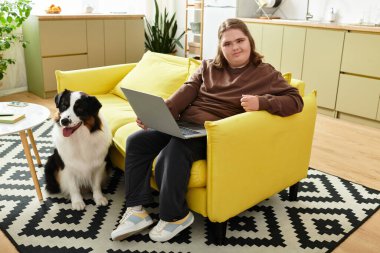 Rahat bir yuva ortamında Down sendromlu genç bir kadın köpeğinin yanında dizüstü bilgisayar kullanırken gülümsüyor..