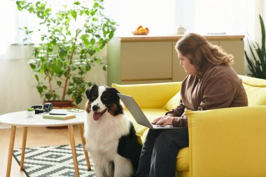 Down sendromlu genç bir kadın sarı bir koltukta oturuyor, yanında köpeğiyle laptopunu kullanıyor..
