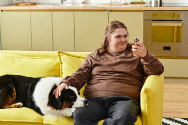 Genç bir kadın evdeki sarı bir kanepede rahatlıyor, telefonunu yanında köpeğiyle mutlu bir şekilde kullanıyor..